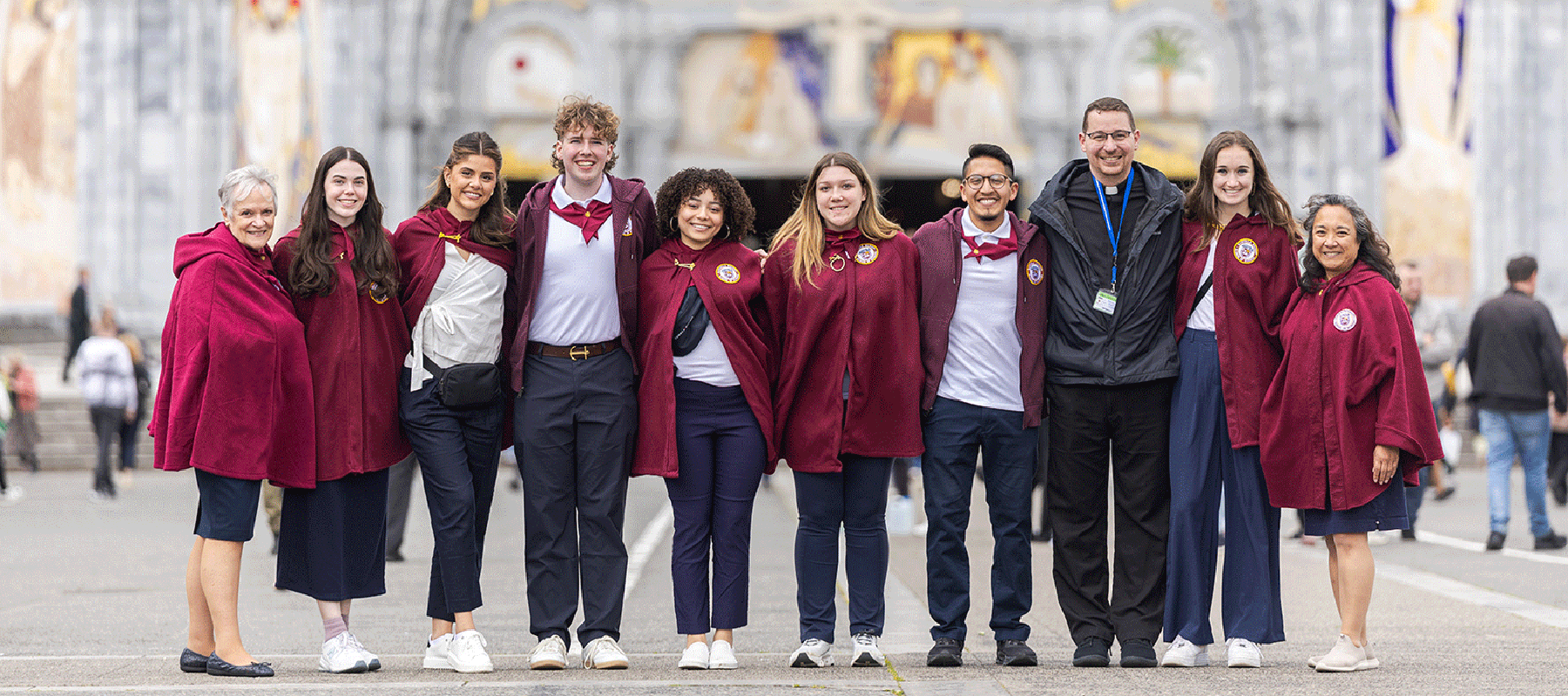 Students in maroon capes stand in line in front of a cathedral, smiling
